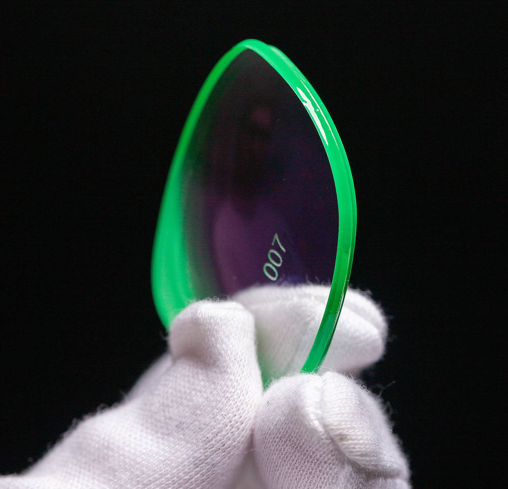 Hand mit Stoffhandschuh praesentiert ein einziges Brillenglas mit shape line Randbeschichtung